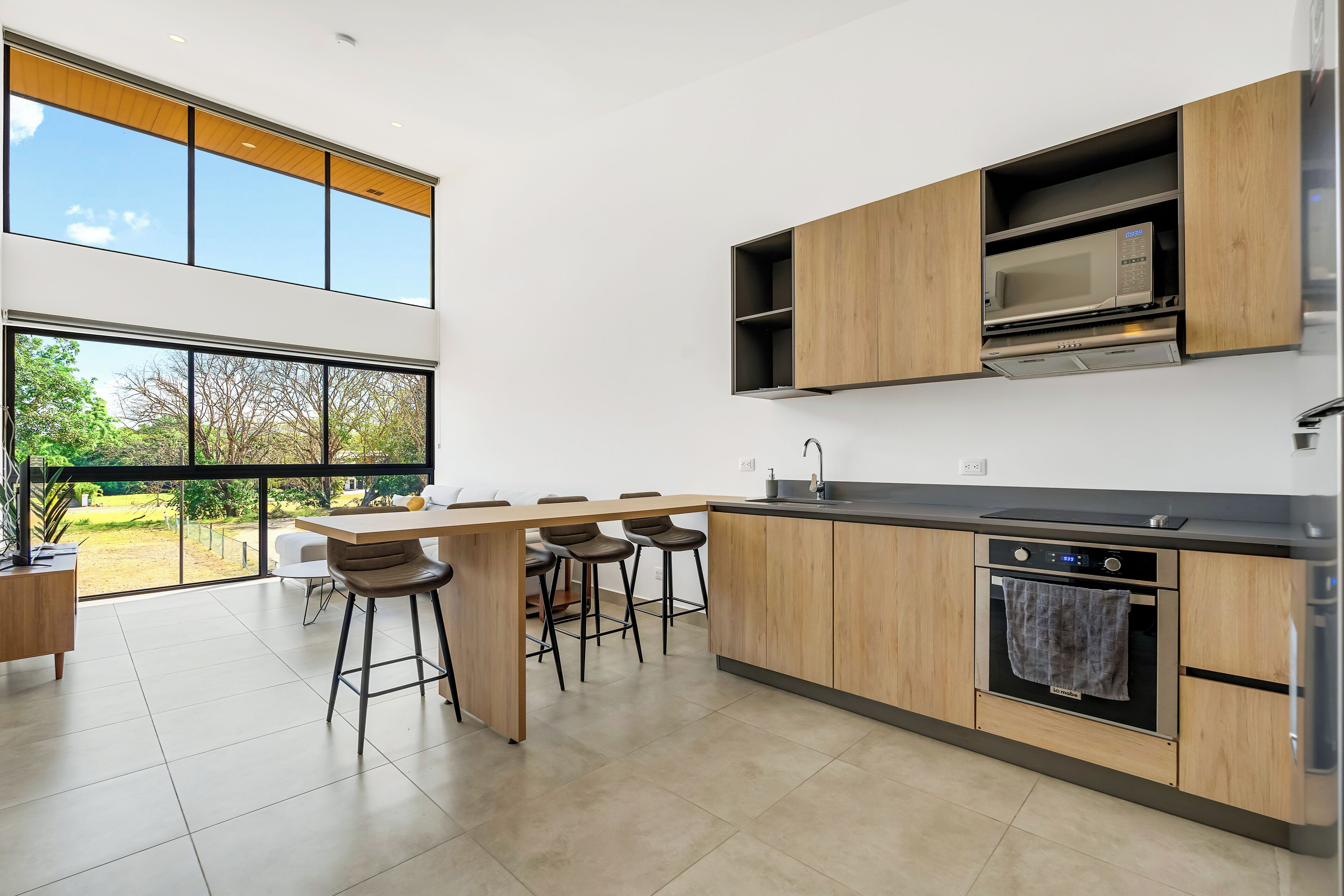 Eine moderne Küche mit großem Fenster, Holzschränken, einem eingebauten Backofen und einer Bar mit Hockern.