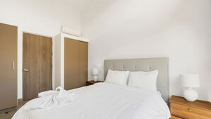 En pænt redt seng med hvide sengetæpper, et trænatbord med en lampe og en dør med et håndklæde hængende over.