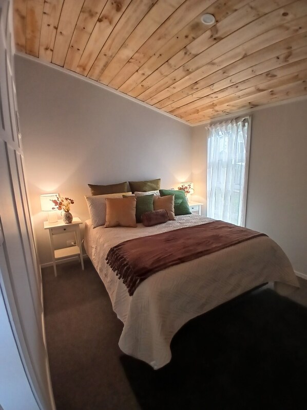 A bedroom with a wooden ceiling, a bed with multiple pillows, a nightstand with a lamp, and a window with curtains.