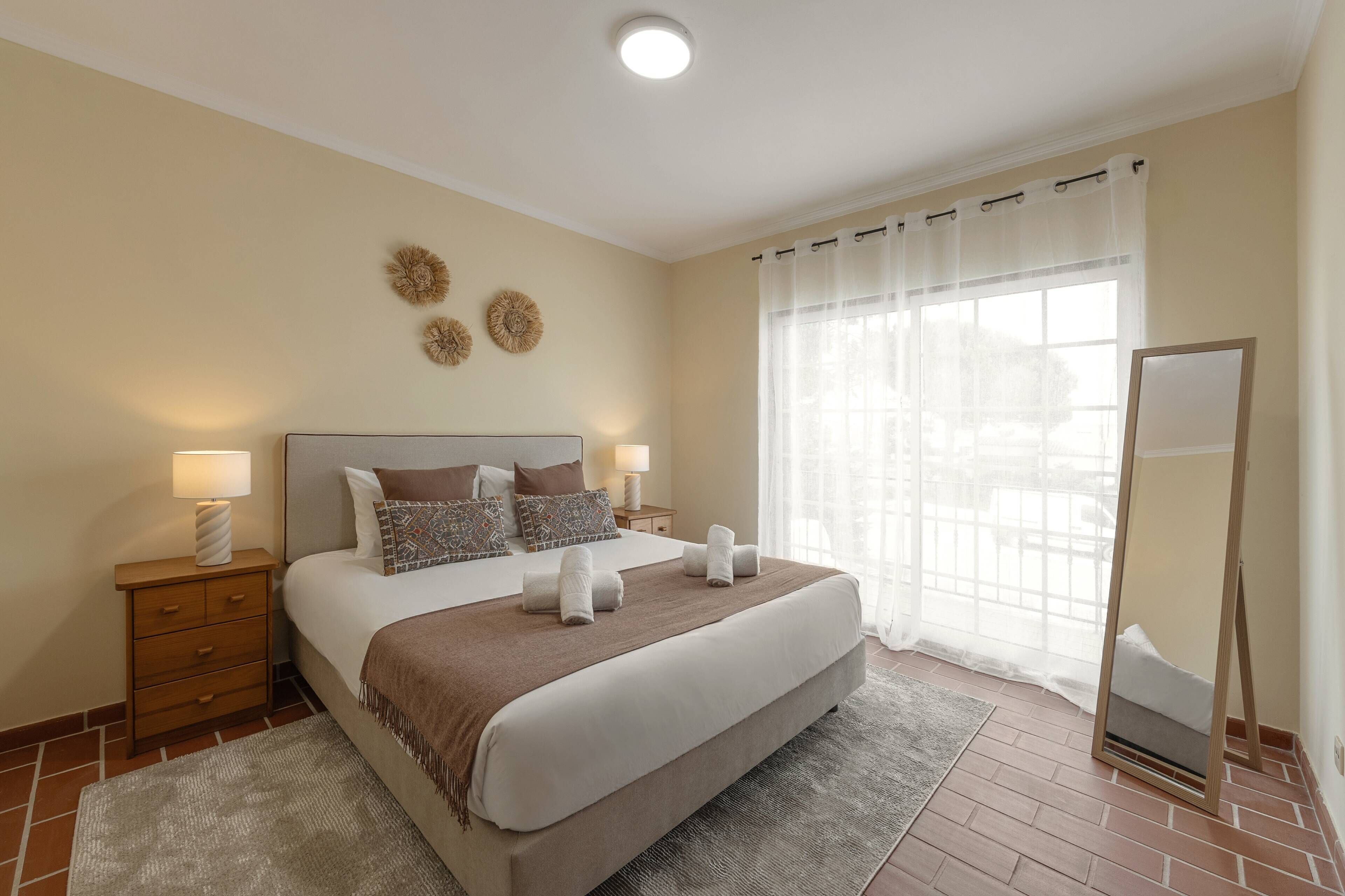 A neatly arranged bedroom with a large bed, bedside tables, a mirror, and a window with sheer curtains.