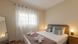A hotel room with a bed, a nightstand, a mirror, a window with sheer curtains, and a brick floor. - Jardim da Lagoa - Rustic Retreat (Óbidos)