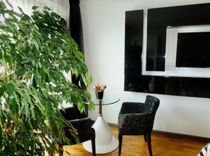 Ein Zimmer mit einer großen Pflanze, einem runden Glastisch, einem gemusterten Sessel und einem Fenster mit Jalousien.