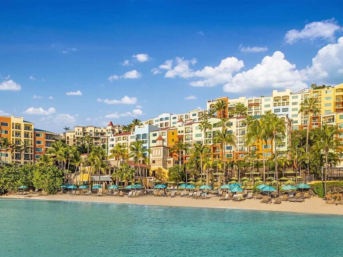 Un hôtel situé en bord de mer, composé de plusieurs bâtiments, agrémenté de palmiers et offrant un accès à une plage équipée de transats.