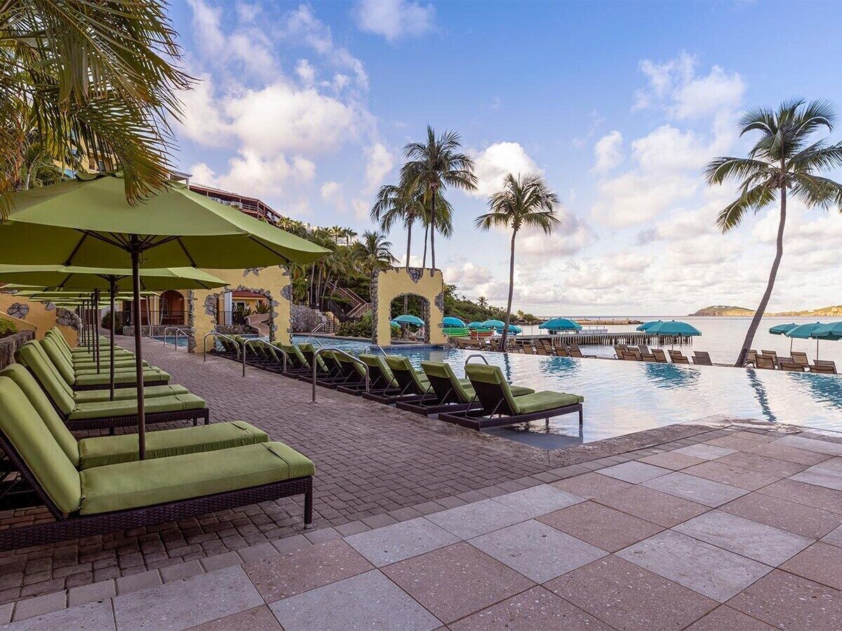 Un espace aménagé au bord de la piscine, avec des chaises longues et des parasols, un ciel dégagé et des palmiers.