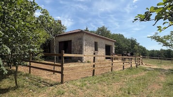 A stone building with a red-tiled roof, surrounded by a wooden fence, and two horses in the background.