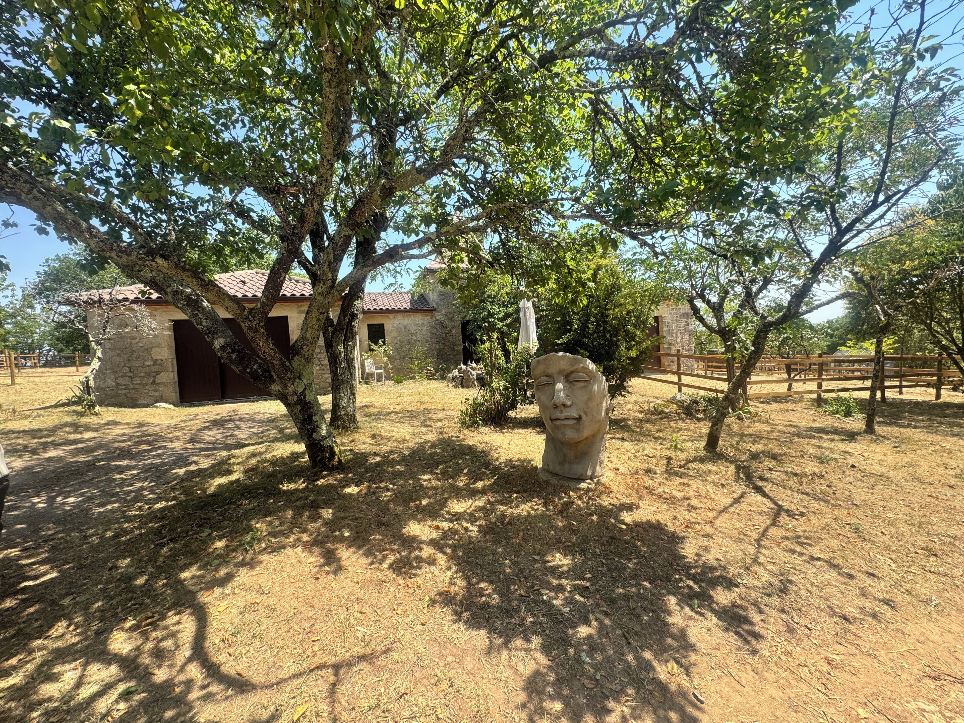 Edificio de piedra con techo de tejas, rodeado de árboles y una zona cubierta de hierba seca.