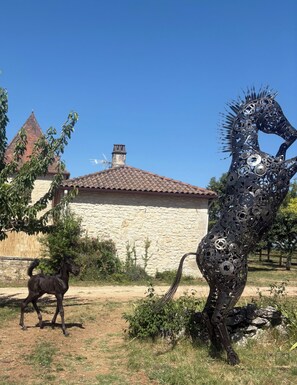 A sculpture of a horse made of metal parts, a black horse, and a stone building with a tiled roof. - Breathtaking stone house with a dovecote near the horses (Mauroux)