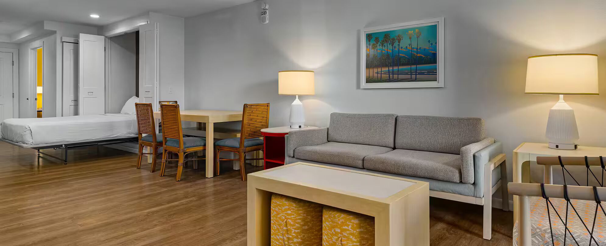 A hotel room with a bed, a grey sofa, a dining table with chairs, two bedside lamps, a painting on the wall, and a wooden floor.