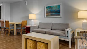 Habitación de hotel con cama, sofá gris, mesa de comedor con sillas, dos lámparas de noche, un cuadro en la pared y suelo de madera.