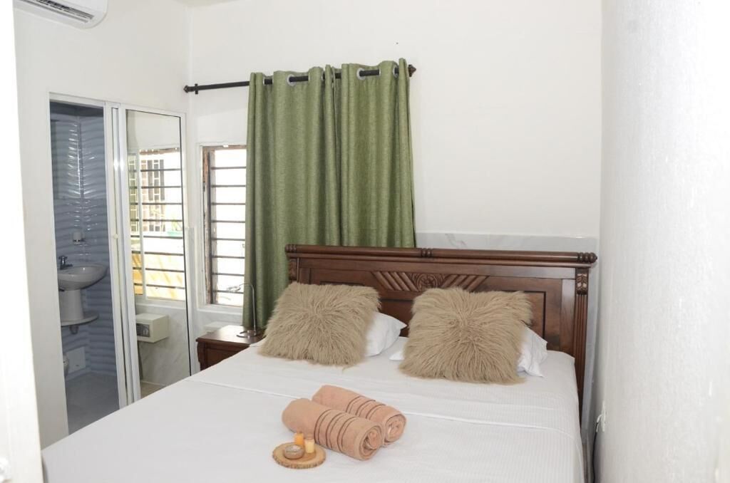 Un dormitorio con cama de madera, dos almohadas mullidas, una cortina verde, una ventana y un baño visible a través de una puerta abierta.