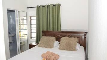A bedroom with a wooden bed, two fluffy pillows, a green curtain, a window, and a bathroom visible through an open door.
