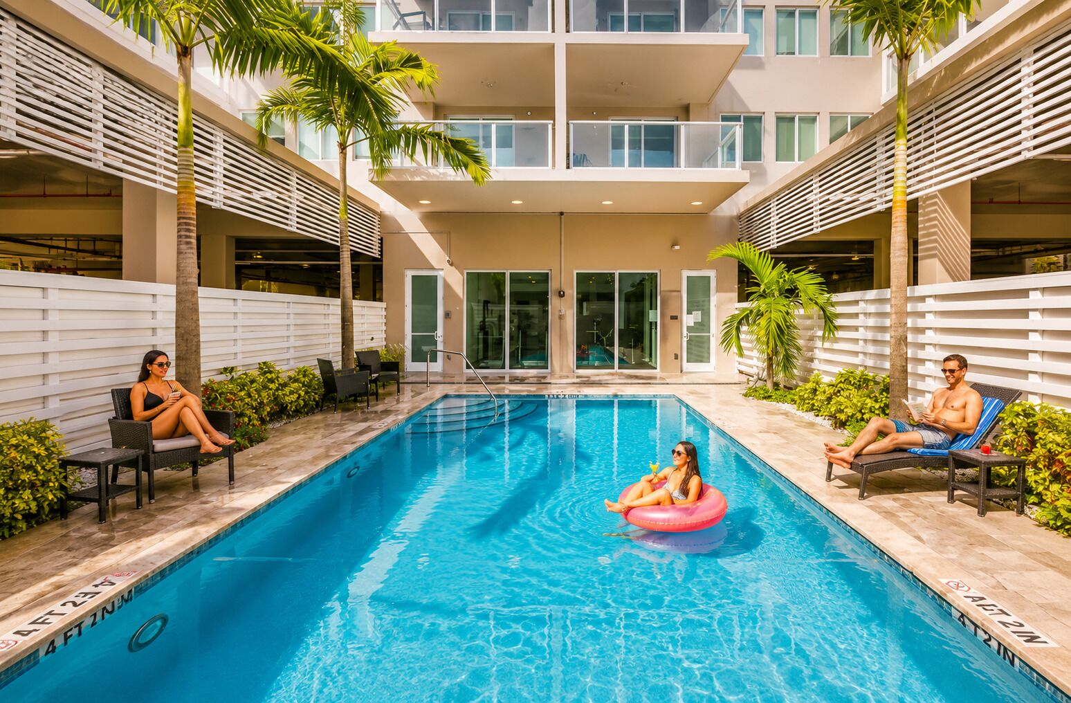 A swimming pool with lounge chairs and palm trees, surrounded by a building with large windows and a white fence.