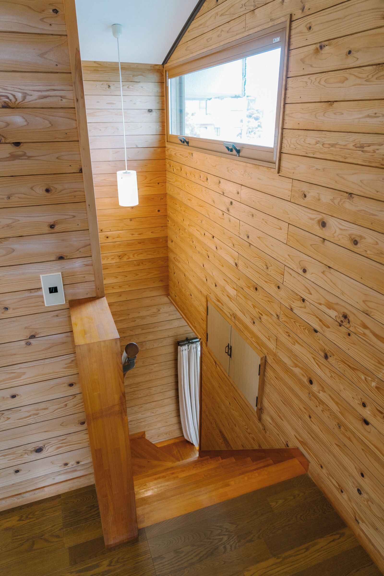 L’intérieur d’une cabane en bois, avec une fenêtre, un luminaire, un banc en bois et une porte.