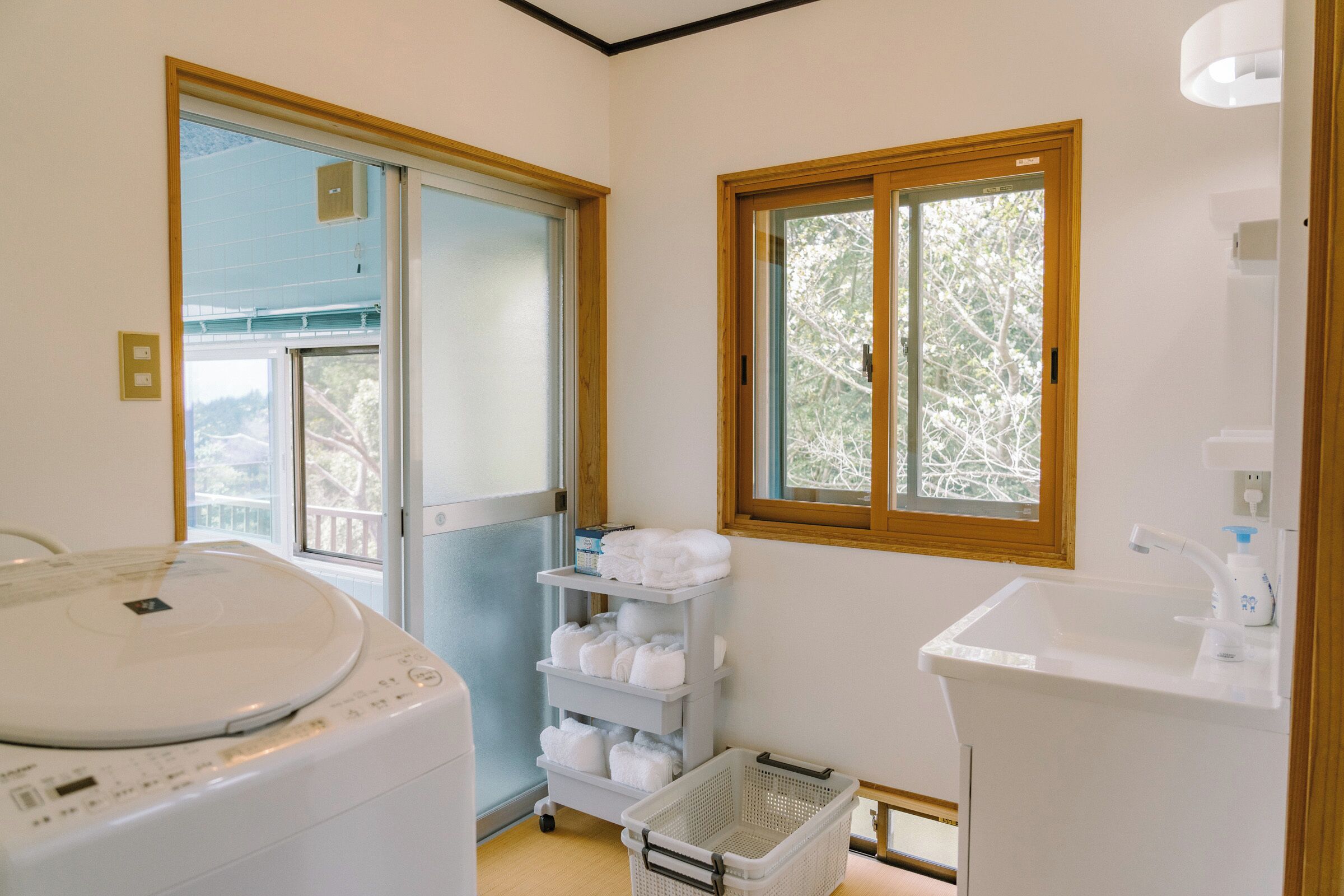 A laundry room with a washing machine, a drying rack, a shelf with folded towels, a basket, and a window with a view of trees.