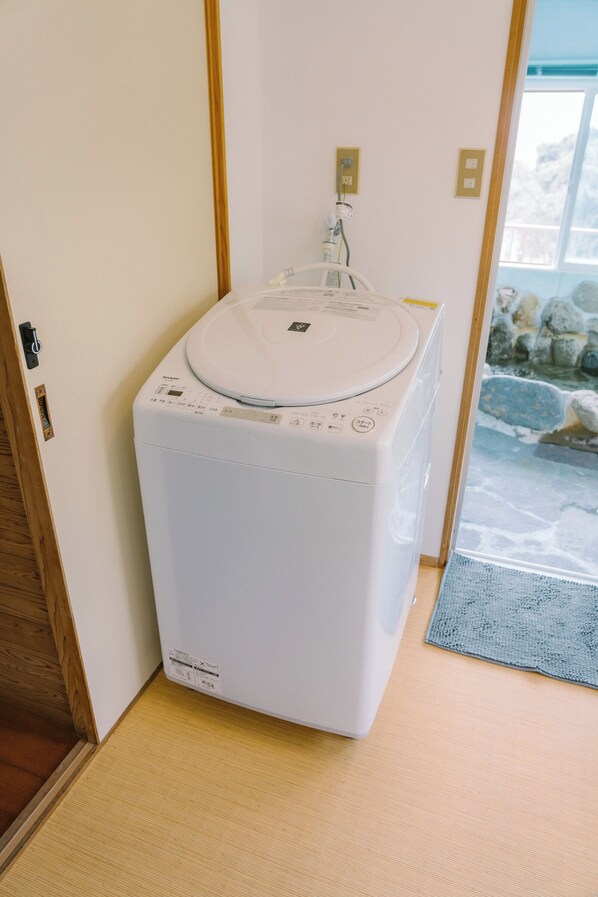 A compact, white washing machine in a corner of a room with a wooden door and a stone wall visible through a window.