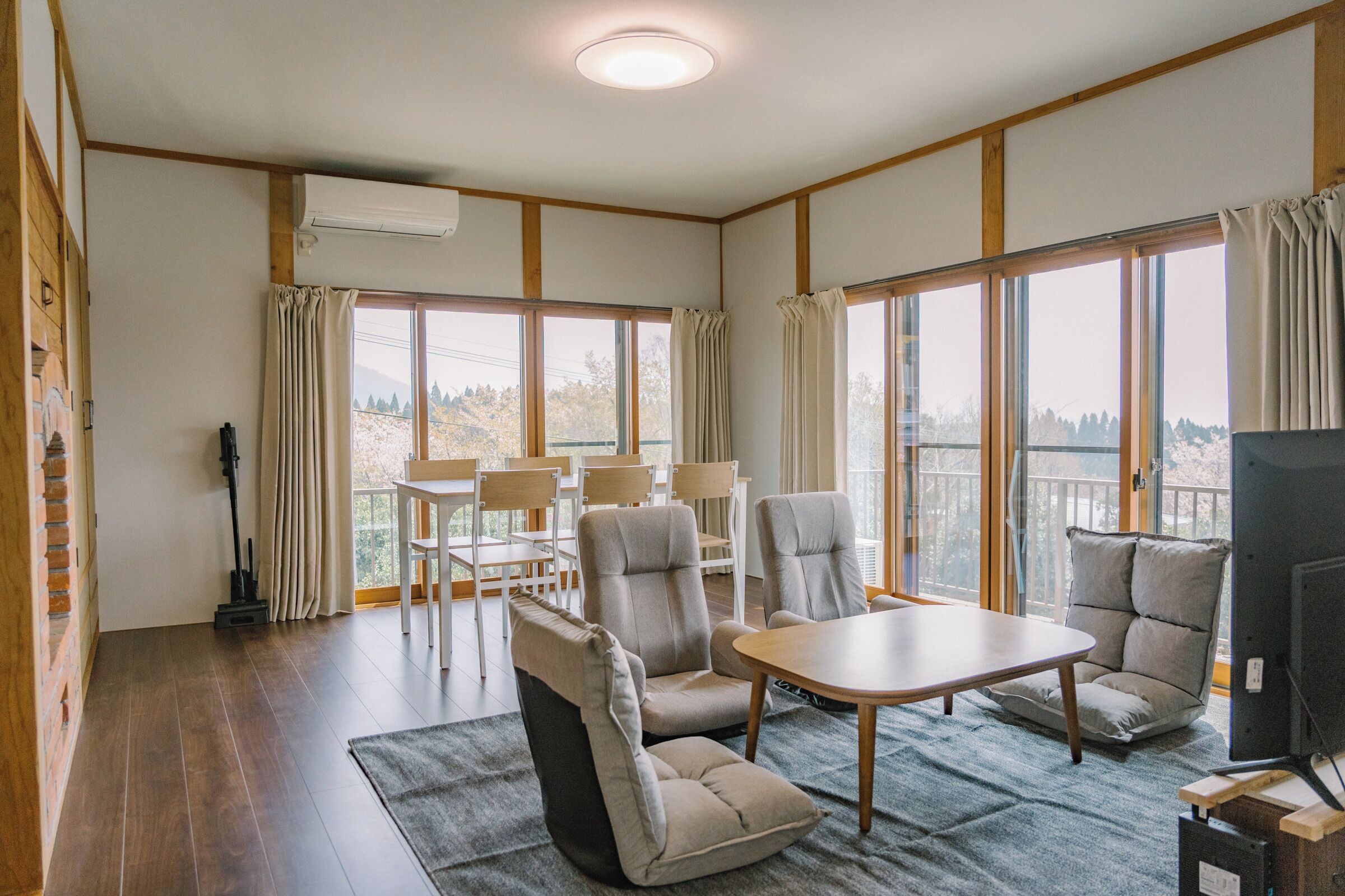 A room with a wooden floor, a table, chairs, a TV, and large windows with curtains.