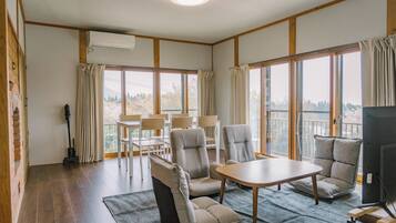 A room with a wooden floor, a table, chairs, a TV, and large windows with curtains.