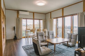 Ein Zimmer mit Holzboden, einem Tisch, Stühlen, einem Fernseher und großen Fenstern mit Vorhängen.