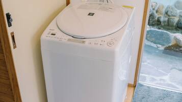 A compact, white washing machine in a corner of a room with a wooden door and a stone wall visible through a window.