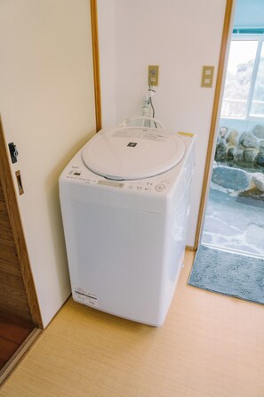 Una lavadora blanca y compacta ubicada en un rincón de una habitación, con una puerta de madera y una pared de piedra visibles a través de una ventana.