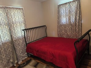 Una cama individual con estructura metálica y colcha roja en una habitación con cortinas estampadas y paredes lisas.