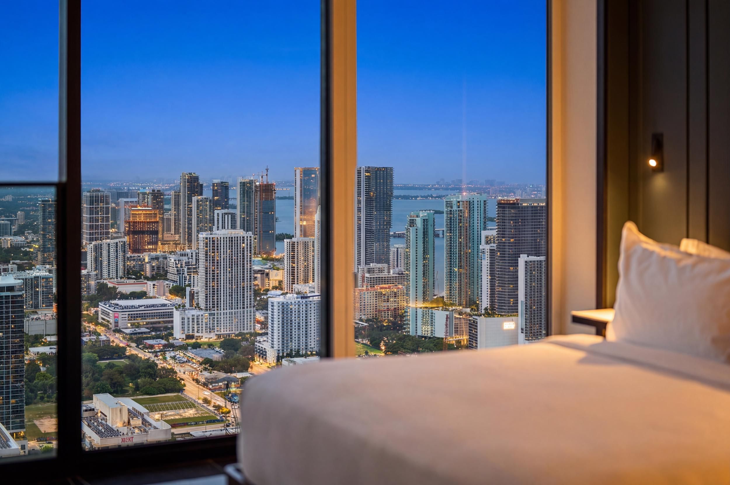 Bed beside city-view windows at dusk.