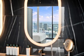A modern hotel room with a large window, a bed, a round mirror, and a bathroom counter with toiletries. - Luxury Sky Penthouse with Ocean & City Views (Miami)