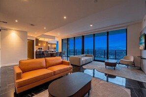 A modern living room with a large sofa, a coffee table, and a view of the city at dusk. - Luxury Sky Penthouse with Ocean & City Views (Miami)