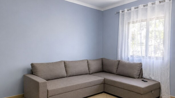 A corner living room with a beige sectional sofa, a window with sheer curtains, and a tiled floor. - A holiday home in a quiet neighborhood 5 minutes walking distance from the sea. (Vlora)
