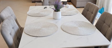 A dining area with a white table, grey upholstered chairs, and a grey sofa in the background.