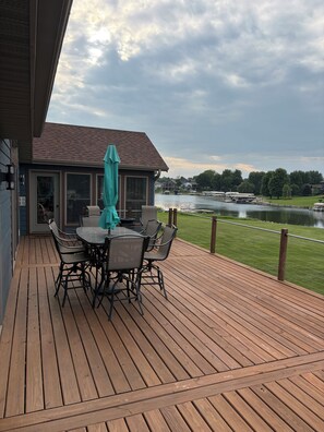 Eine Holzterrasse mit Tisch und Stühlen, mit Blick auf ein Gewässer und eine Rasenfläche.