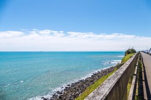 A coastal road with a concrete barrier, rocky shoreline, and the ocean. - Waterfront On Woolcombe - Spacious Holiday Home (New Plymouth)