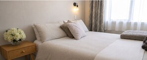A neatly made bed with a striped bedspread and pillows, a bedside table with a vase of flowers, and a wall-mounted light fixture. - Waterfront On Woolcombe - Spacious Holiday Home (New Plymouth)