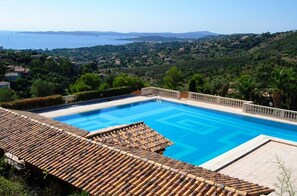 Ein Schwimmbecken mit einer mit Fliesen ausgelegten Terrasse, umgeben von Grünflächen und Blick auf eine entfernte Küstenlinie.