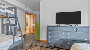 A bedroom with a bunk bed, a dresser, a television, and a door leading to another room. - SALTED SHORES OBX formerlyPLAYING HOOKY, 5bed,Wild Horses,Pool,Hot Tub,4x4 Beach (Corolla)