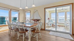 A dining area with a wooden table and white chairs, a sailboat decoration, and a view through a glass door to an outdoor balcony. - SALTED SHORES OBX formerlyPLAYING HOOKY, 5bed,Wild Horses,Pool,Hot Tub,4x4 Beach (Corolla)