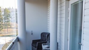 Un balcón con una silla de mimbre, una mesita pequeña, una puerta blanca y vistas a la calle y árboles.