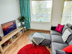 Ein Wohnzimmer mit Fernseher, einer Couch, einem roten Teppich und einem Fenster mit Jalousien.