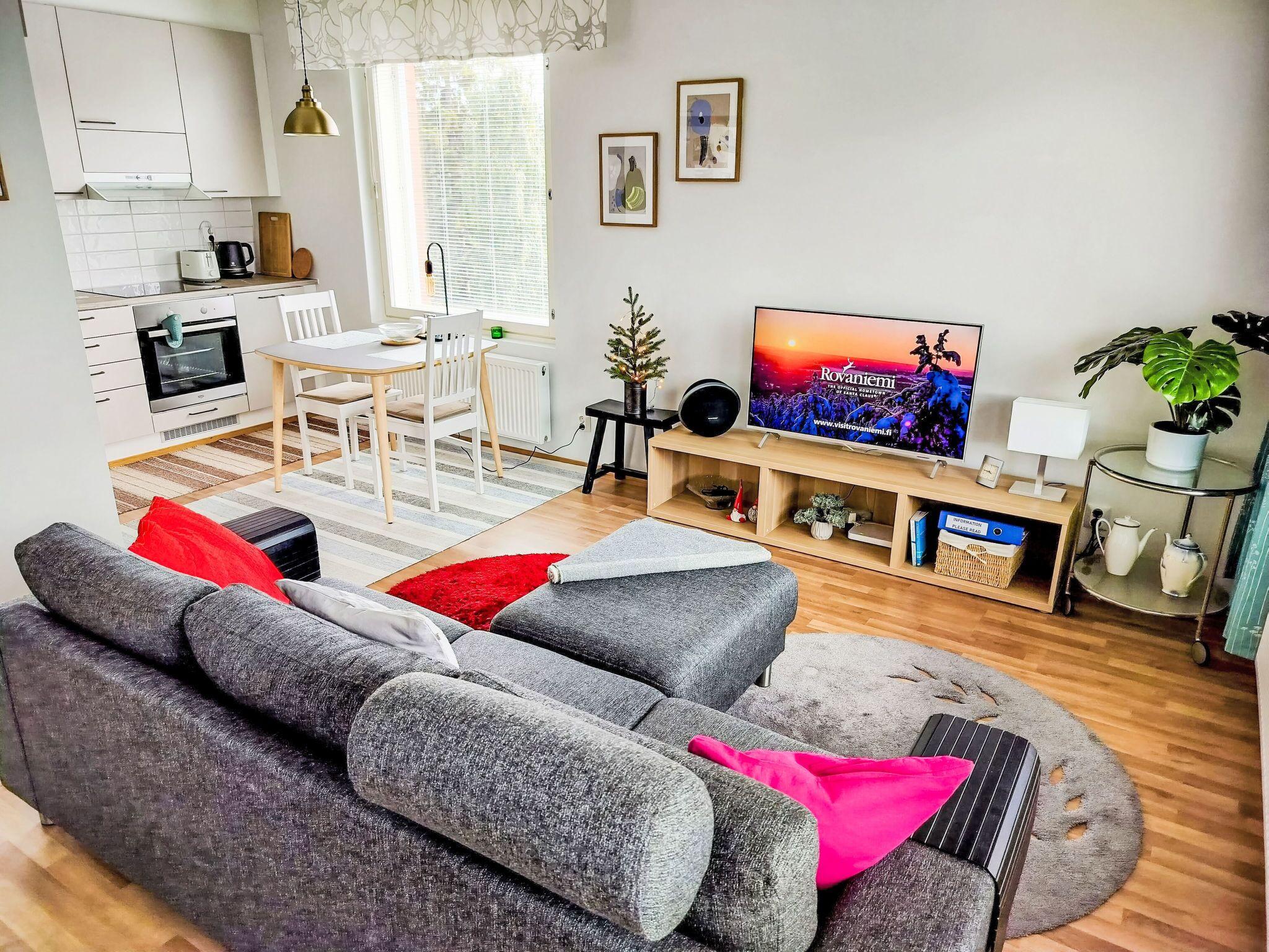 A modern living room with a grey sofa, a dining table, a TV, and a small Christmas tree.