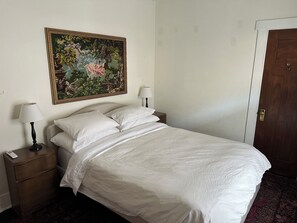 Una cama bien hecha con sábanas blancas, una mesita de noche con lámpara, un cuadro en la pared y una puerta a la derecha.