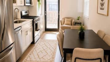 Une cuisine moderne équipée d’appareils électroménagers en acier inoxydable, un coin repas avec une table sombre et des chaises beiges, et une vue à travers une porte vitrée.
