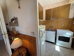 Eine Küche mit Herd, Spüle und weißen Schränken. Es gibt einen Holzkasten mit einer Ablage, eine Lampe und einen kleinen Tisch mit einem Bilderrahmen.