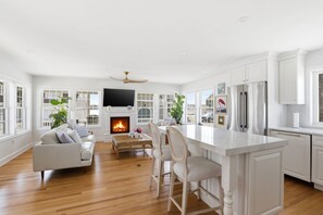 A spacious living room with a fireplace, a dining area with bar stools, and a kitchen with white cabinetry. - Newly renovated 3BR/2BA coastal beach retreat-sweeping harbor & ocean views. (Scituate)