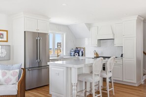 A modern kitchen with white cabinetry, a stainless steel refrigerator, and a central island with bar stools. - Newly renovated 3BR/2BA coastal beach retreat-sweeping harbor & ocean views. (Scituate)