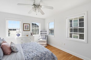 Une chambre à coucher avec un lit, une commode, une chaise et de grandes fenêtres.