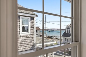 A window view of a coastal house with a wooden exterior, a white window frame, and a clear view of the sea and other houses. - Newly renovated 3BR/2BA coastal beach retreat-sweeping harbor & ocean views. (Scituate)
