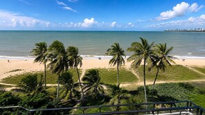 A beach with palm trees, a clear sky, and a distant city skyline. - Beachfront property in Bessa | Ocean view with pool (João Pessoa)