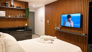 Habitación de hotel con cama, televisor montado en la pared, paneles de madera, una zona de cocina con lavabo y una planta.