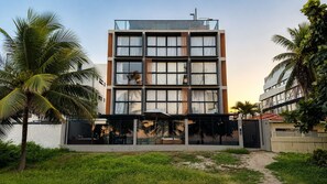 A multi-story building with large windows, a covered outdoor seating area, and palm trees. - Beachfront property in Bessa | Ocean view with pool (João Pessoa)