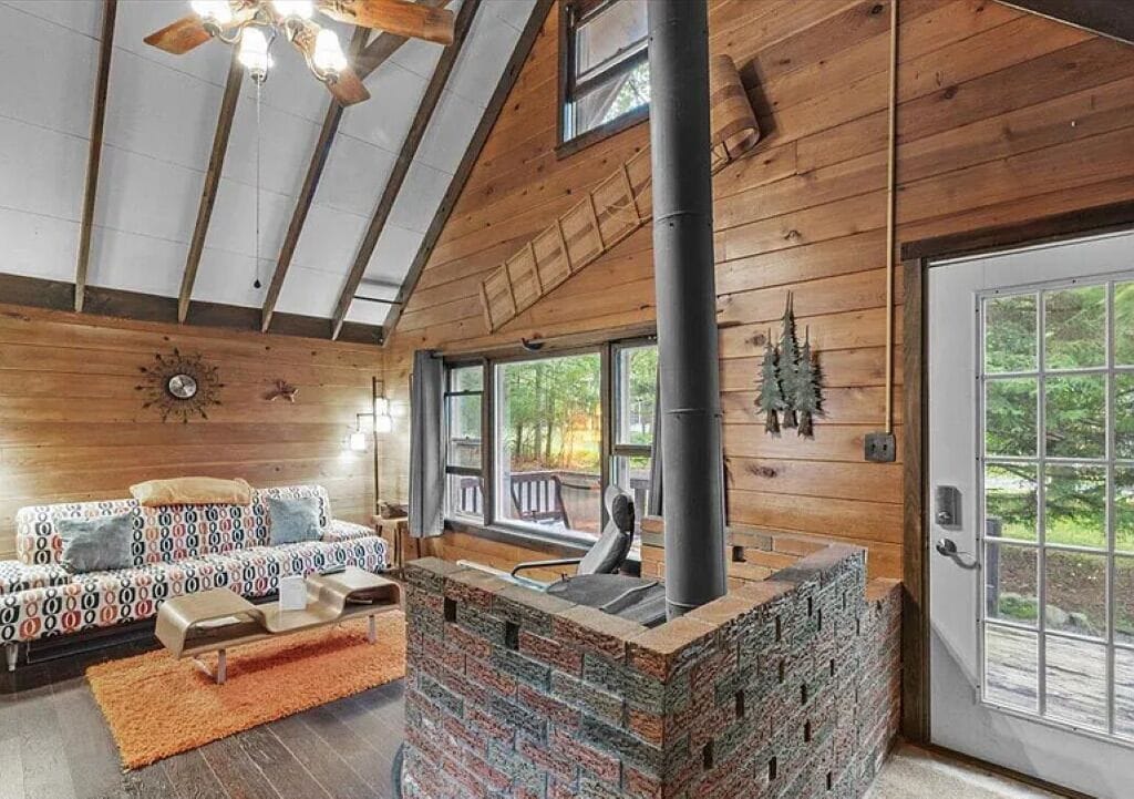 A log cabin interior with a fireplace, wooden walls, a sofa, and a view through large windows.